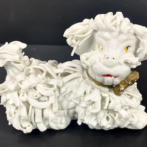 May include: White porcelain figurine of a dog with a curly, shaggy coat. The dog has a painted black nose, red mouth, and yellow eyes. The dog is wearing a gold collar with a brown bow.