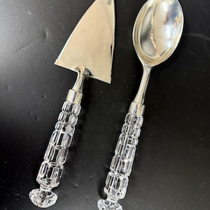 May include: A silver cake server and serving spoon set. The serving pieces have clear, faceted crystal handles with silver accents. The server has a triangular blade, and the spoon has a rounded bowl. The set is displayed on a black surface.