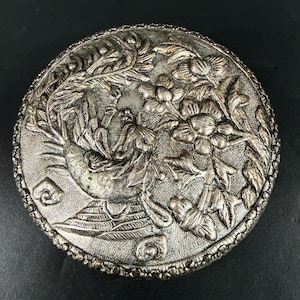 May include: A round, silver-toned decorative box with an embossed design. The lid features a detailed relief of a bird, flowers, and foliage. The box has a beaded edge and is set against a dark background.