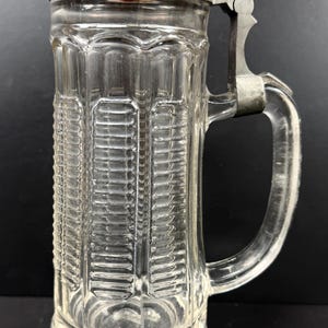 May include: Clear glass beer stein with a hinged metal lid and handle. The stein has a rectangular shape with vertical textured detailing. The lid is silver-colored with a dark red center. The handle is clear glass, and the stein is on a black background.