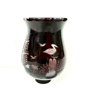 May include: A dark burgundy glass vase with a flared top and a round base. The vase features etched designs of birds, plants, and water elements. The overall design is a nature-themed decorative piece.