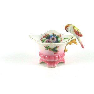 May include: A small, pink and white porcelain cup with a floral design and a gold-painted handle. A yellow and orange bird is perched on the handle.