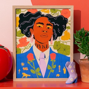 以下が含まれることがあります： ダークヘアの人物の肖像画が描かれた額入りアートプリント。青いブレザー、白いシャツ、ネクタイを着用しており、首元には「DYKE」の文字がある。背景にはオレンジや黄色の花が描かれ、馬や魚の骨などの小さなアイコンが散りばめられている。