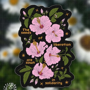 May include: A die-cut sticker featuring pink hibiscus flowers with green leaves on a black background. The text reads: The seeds of liberation are sown in solidarity. Small yellow stars are scattered throughout the floral illustration.