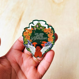 Puede incluir: Una pegatina verde con una corona de flores y un querubín con alas. La corona está adornada con flores naranjas, flores moradas y hojas verdes. El querubín tiene piel morena y sus manos están en sus mejillas. La pegatina dice "Live, Laugh, Love for Liberation."