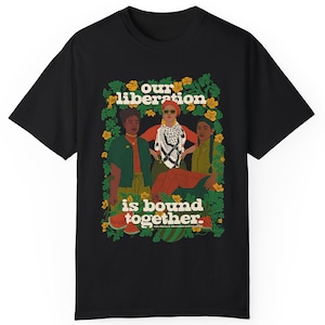 Puede incluir: Camiseta negra con un estampado gráfico de tres personas de pie juntas frente a un fondo verde frondoso. El texto "our liberation is bound together." está impreso debajo de las personas.