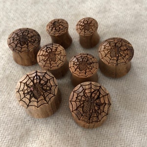 May include: A collection of seven wooden ear plugs. Each plug features a detailed spider web design with a spider in the center. The plugs are made of light brown wood and are suitable for stretched ear piercings.