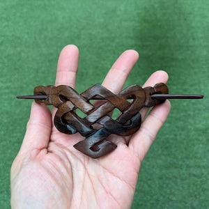 May include: A dark brown wooden hair barrette with an intricate Celtic knot design. The barrette features a long, slender pin for securing the hair. The wood grain is visible, adding to the handcrafted aesthetic.