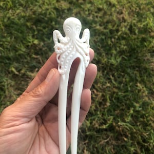 May include: Ivory-colored hair fork with an octopus design. The octopus is carved at the top of the fork, with its tentacles and head detailed. The fork has two long, smooth prongs. The background is green grass.