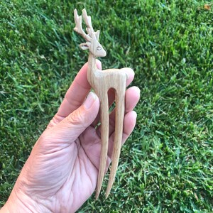 May include: A light brown wooden hair fork carved in the shape of a deer. The deer has detailed antlers and a slender body. The hair accessory is held against a background of green grass.