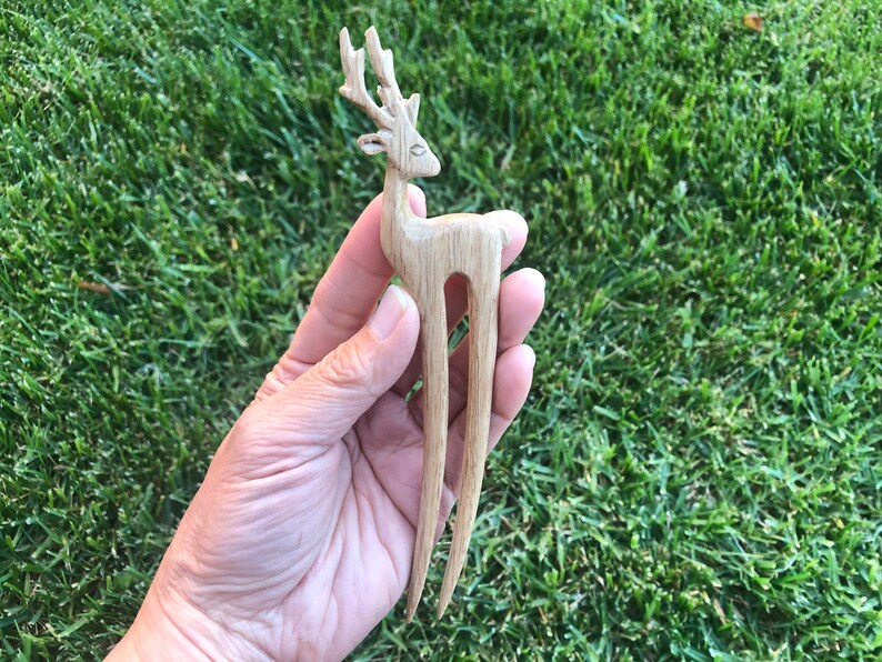 May include: A light brown wooden hair fork carved in the shape of a deer. The deer has detailed antlers and a slender body. The hair accessory is held against a background of green grass.