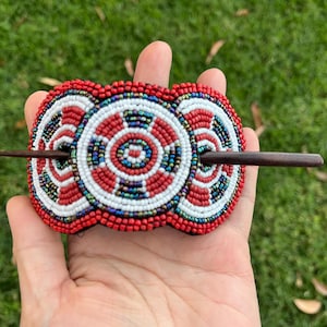 Op de afbeelding: Een met kralen versierde haarspeld met een houten stok. De haarspeld heeft een cirkelvormig ontwerp met rode, witte en blauwe kralen. De haarspeld wordt in een hand gehouden tegen een groene achtergrond.