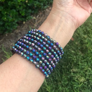 May include: A stack of iridescent bracelets made of small, round beads. The beads are a dark purple color with a rainbow sheen.