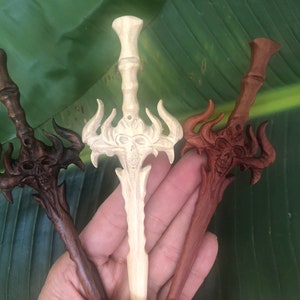 May include: Three wooden hair sticks carved in the shape of swords with skull details. The swords are in natural, dark brown, and reddish brown wood.