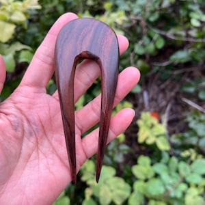 May include: A dark brown wooden hair fork with a U-shaped top and two long, pointed tines. The hair accessory is held in a hand against a backdrop of green foliage.