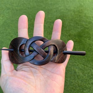 May include: Dark brown wooden hair barrette with an intricate Celtic knot design. The barrette features a long, thin pin for securing the hair. The wood grain is visible, adding to the natural aesthetic. A stylish hair accessory.