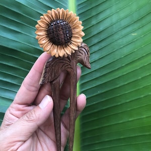 Può includere: Un fermaglio per capelli in legno con un girasole intagliato sulla parte superiore, caratterizzato da un centro marrone scuro e petali marrone chiaro. Il gambo del fermaglio è diviso in due punte, con un dettaglio a foglia alla base.