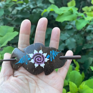 May include: A brown wooden hair barrette with a decorative design. The barrette features a white flower with a purple center and blue accents. A wooden stick is inserted through the barrette to secure it in the hair.