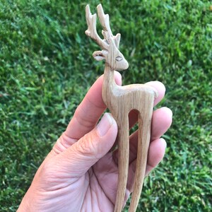 May include: A wooden hair fork carved in the shape of a deer. The deer has detailed antlers and a slender body. The hair accessory is made of light-colored wood and is held against a background of green grass.