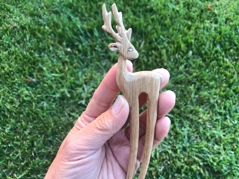 May include: A wooden hair fork carved in the shape of a deer. The deer has detailed antlers and a slender body. The hair accessory is made of light-colored wood and is held against a background of green grass.