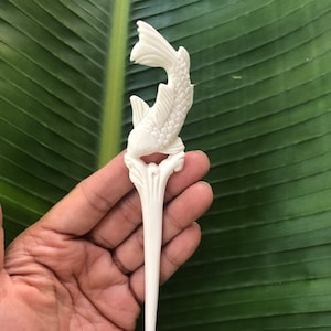 May include: Ivory-colored, carved fish hair stick. The fish is depicted in a stylized, upward-curving pose, with detailed scales and fins. The stick has a floral base and a long, pointed shaft. The background is a large green leaf.