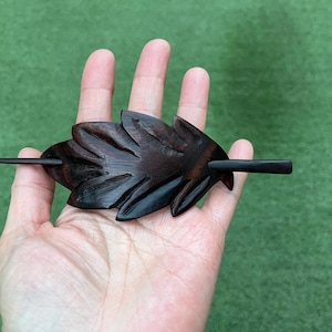 Op de afbeelding: Een donkerbruine houten haarspeld in de vorm van een blad. Het haaraccessoire heeft een lange, dunne pin om het in het haar te bevestigen. Het bladerontwerp heeft gedetailleerde gesneden lijnen en het hout heeft een gepolijste afwerking.