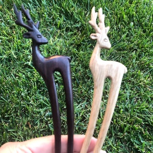 May include: Two wooden hair forks, each featuring a deer design. One is dark brown, the other is light wood. The deer have detailed antlers and a curved body, with a long, slender fork extending from the base.