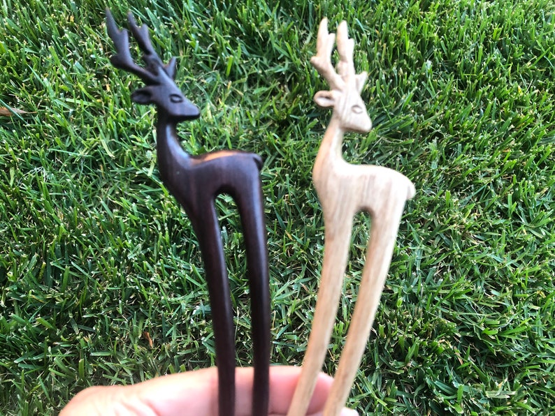 May include: Two wooden hair forks, each featuring a deer design. One is dark brown, the other is light wood. The deer have detailed antlers and a curved body, with a long, slender fork extending from the base.
