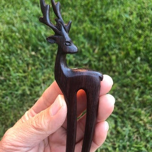 May include: A dark brown wooden hair fork carved in the shape of a deer. The deer has detailed antlers and a slender body. The fork is held against a background of green grass.