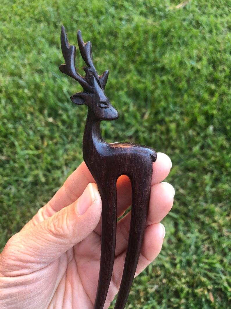 May include: A dark brown wooden hair fork carved in the shape of a deer. The deer has detailed antlers and a slender body. The fork is held against a background of green grass.