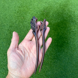 May include: Two dark brown wooden hair sticks with leaf and branch designs. The hair accessories are carved with intricate details and held in a hand against a green background.