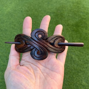 May include: Dark brown wooden hair barrette with an intricate, swirling design. The barrette features a central decorative element with a pin for securing it in the hair. The hair accessory is held in a hand.