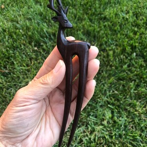 May include: A dark brown wooden hair fork featuring a deer design. The deer has detailed antlers and a slender body. The fork has two long, pointed tines, perfect for securing hair in an updo. The hair accessory is held against a backdrop of green grass.
