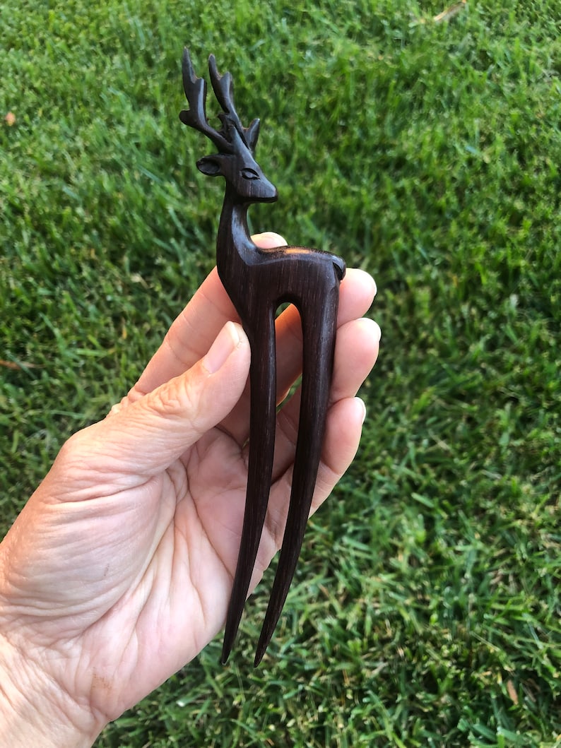 May include: A dark brown wooden hair fork featuring a deer design. The deer has detailed antlers and a slender body. The fork has two long, pointed tines, perfect for securing hair in an updo. The hair accessory is held against a backdrop of green grass.