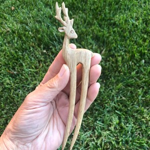 May include: A wooden hair fork featuring a carved deer design. The deer has detailed antlers and a slender body. The fork's tines are long and pointed. The hair accessory is made of light-colored wood.