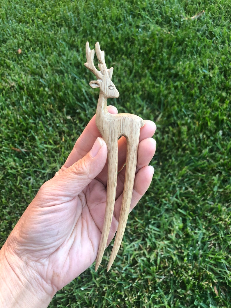 May include: A wooden hair fork featuring a carved deer design. The deer has detailed antlers and a slender body. The fork's tines are long and pointed. The hair accessory is made of light-colored wood.