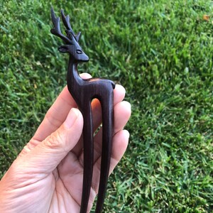 May include: A dark brown wooden hair stick with a deer head design. The deer has detailed antlers and a slender body. The hair accessory is held against a backdrop of green grass.