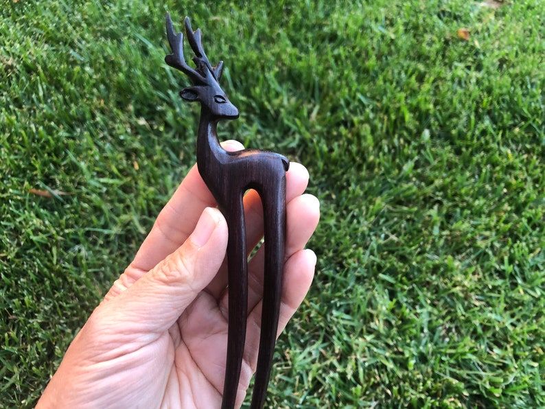 May include: A dark brown wooden hair stick with a deer head design. The deer has detailed antlers and a slender body. The hair accessory is held against a backdrop of green grass.