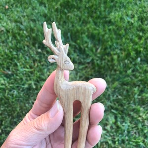 May include: A wooden hair fork featuring a carved deer design. The deer has detailed antlers and a slender body. The hair accessory is made of light-colored wood and is held against a background of green grass.