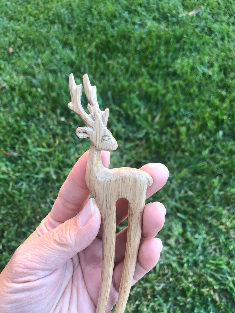 May include: A wooden hair fork featuring a carved deer design. The deer has detailed antlers and a slender body. The hair accessory is made of light-colored wood and is held against a background of green grass.