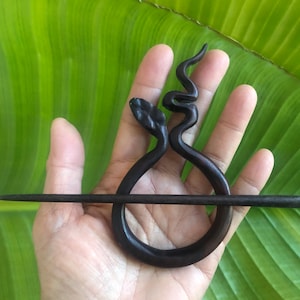 May include: Dark brown wooden hair pin featuring a stylized snake design. The pin has a circular loop with a snake head and a curved body. A straight, dark brown stick goes through the loop. The background is a large green leaf.