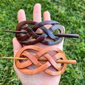 May include: Two wooden hair sticks in a Celtic knot design. One is dark brown, the other is light brown. Each has a wooden pin through the center. The hair accessories are held in a hand against a green background.