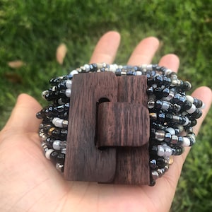 Op de afbeelding: Een kralenarmband met een donkere houten sluiting. De armband heeft een mix van zwarte, witte en zilveren kralen. De sluiting is rechthoekig met een gebogen sluiting. De armband wordt in een hand gehouden tegen een groene achtergrond.