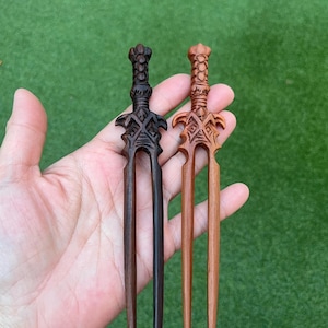 May include: Two wooden hair forks, each with a decorative sword-like design. One is dark brown, the other is light brown. The forks are held against a green background.