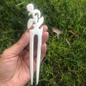 May include: White bone hair stick with a carved floral design. The stick has two prongs and a decorative top.