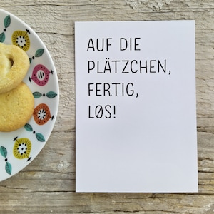 May include: A white rectangular card with black text that reads AUF DIE PL&Auml;TZCHEN, FERTIG, L&Oslash;S! positioned next to a white plate featuring a colorful pattern of stylized yellow, pink, and orange floral shapes with two round sugar cookies.