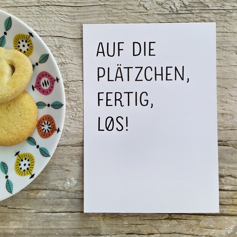May include: A white rectangular card with black text that reads AUF DIE PL&Auml;TZCHEN, FERTIG, L&Oslash;S! positioned next to a white plate featuring a colorful pattern of stylized yellow, pink, and orange floral shapes with two round sugar cookies.