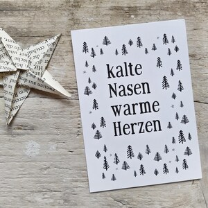 May include: A rectangular white greeting card featuring the black printed text "kalte Nasen warme Herzen" centered on the front. The text is surrounded by a pattern of small black pine tree illustrations and tiny gray snowflake symbols.