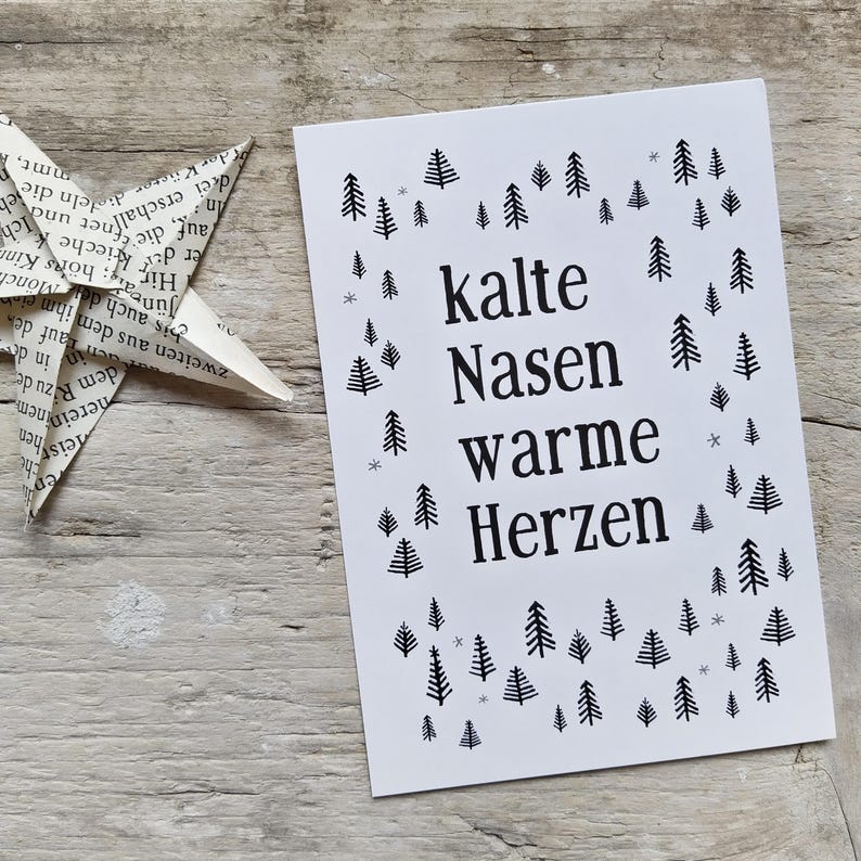 May include: A rectangular white greeting card featuring the black printed text "kalte Nasen warme Herzen" centered on the front. The text is surrounded by a pattern of small black pine tree illustrations and tiny gray snowflake symbols.