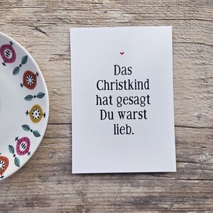 May include: A white rectangular greeting card with black text that reads Das Christkind hat gesagt Du warst lieb and a small red heart icon above the text, positioned next to a white plate with a colorful geometric pattern.